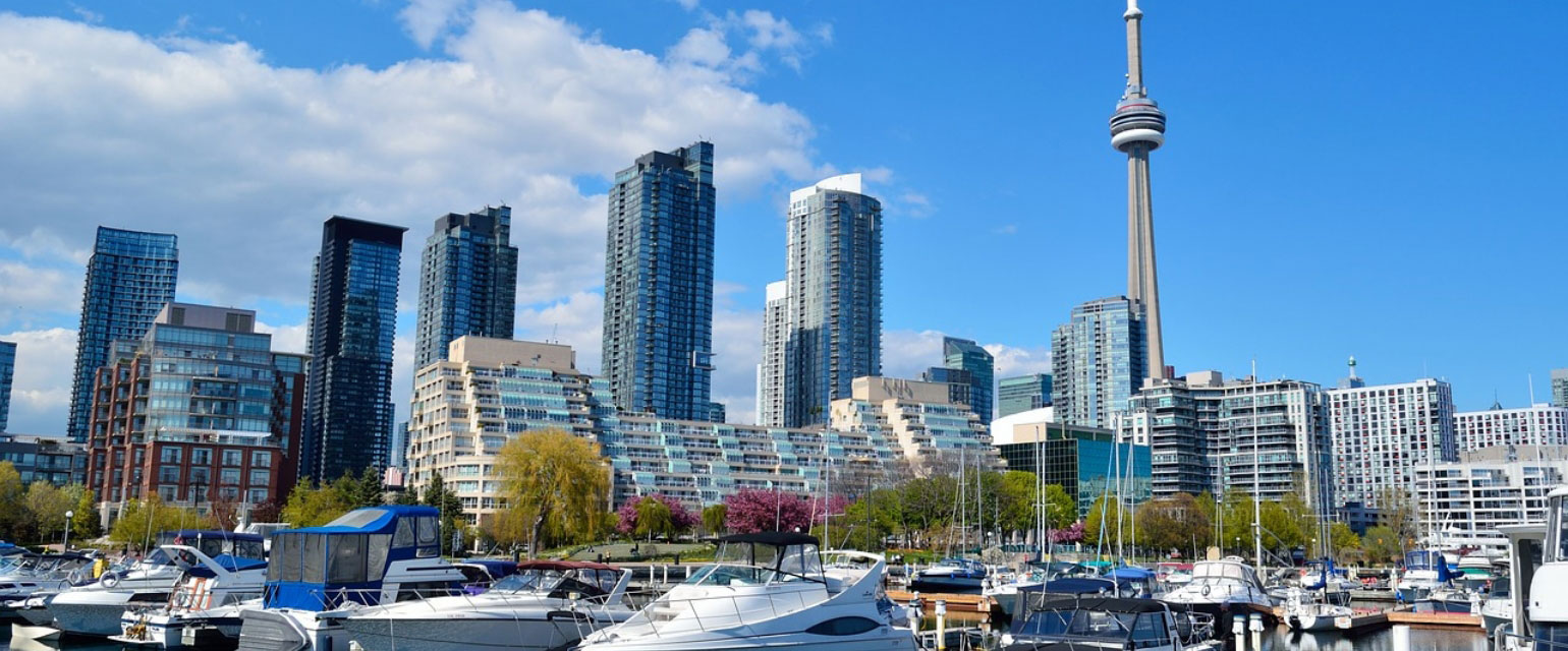 Panoramic blue skies over Toronto landmarks, yachts and motor boats on the pier, luxury waterfront condominiums and the iconic spire of CN Tower next to the calm shore of Lake Ontario, Canada.
